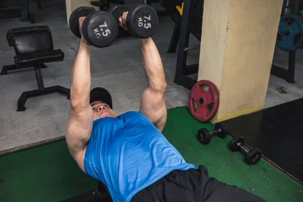 An asian man in a blue tank top does a set of dumbbell chest flys on a flat bench. Training pecs and upper body at the gym. Starting position.