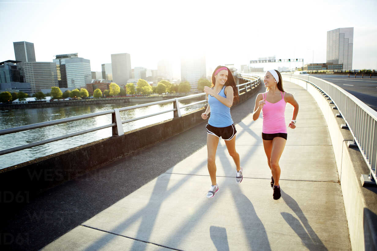 Female athlete friends talking while running on bridge against sky in city during sunny day